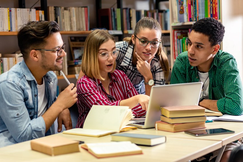 Four students working together around a laptop and books in a library